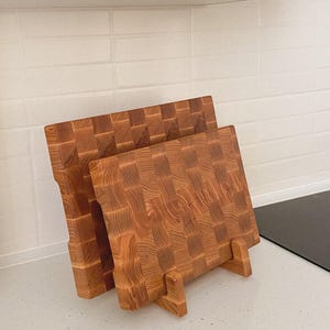 May include: Two wooden butcher block cutting boards with a checkerboard pattern. The boards are made of light brown wood and are displayed upright on a countertop. The boards are supported by small wooden stands.