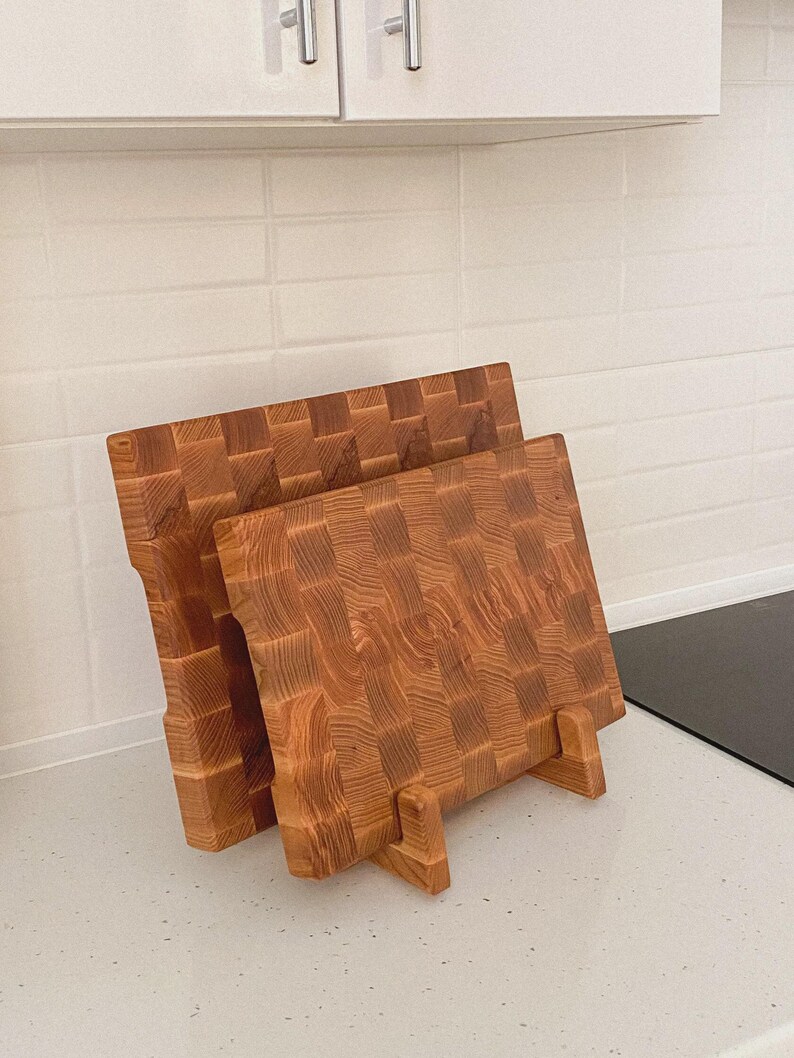 May include: Two wooden butcher block cutting boards with a checkerboard pattern. The boards are made of light brown wood and are displayed upright on a countertop. The boards are supported by small wooden stands.