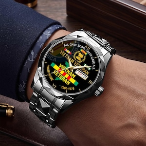 Può includere: Un orologio da polso in acciaio inossidabile nero con lunetta sfaccettata e cinturino a maglie. Il quadrante nero riporta il testo Vietnam Veteran, gli anni 1960-1975 e la frase All Gave Some, 58479 Gave All. Include lancette dorate e data.