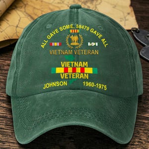 Pode incluir: Um boné de beisebol verde-escuro com texto amarelo que diz ALL GAVE SOME, 58479 GAVE ALL, VIETNAM VETERAN, JOHNSON e 1960-1975. O design inclui um emblema central com três figuras, fitas militares e uma silhueta de mapa verde.