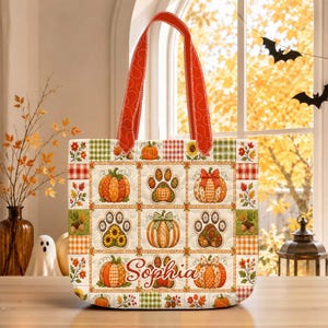 May include: A quilted tote bag featuring a patchwork design of pumpkins, paw prints, and autumn leaves in orange, green, and brown tones. The bag has red patterned handles and the name Sophia written in cursive script across the bottom center.