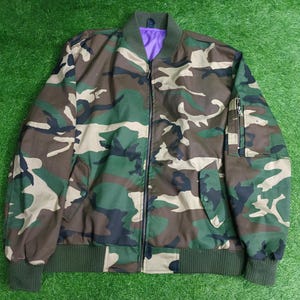 May include: Camouflage bomber jacket with a green ribbed collar, cuffs, and hem. The jacket features a zipper closure, two front pockets, and a zippered pocket on the left sleeve. The interior lining is purple.