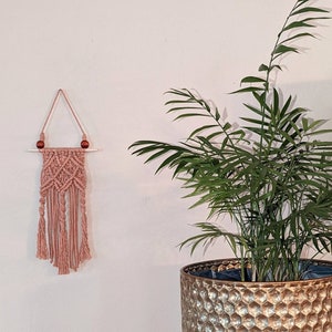 May include: A dusty rose macrame wall hanging with a geometric design and fringe. The wall decor features a wooden dowel and two wooden beads. A potted plant with green leaves is in the background.