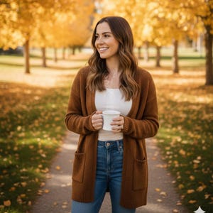 Pode incluir: Uma mulher vestindo um cardigan marrom, top branco e jeans azul, segurando uma caneca branca. O cardigan tem dois bolsos frontais. O fundo apresenta um caminho com árvores e folhas amarelas, sugerindo o outono.