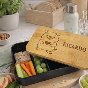 Könnte beinhalten: Eine schwarze Lunchbox mit Bambusdeckel, gefüllt mit Gemüse und Crackern. Der Deckel ist mit einem Bären und dem Namen "Ricardo" graviert. Eine Wasserflasche, Brot und Obst sind ebenfalls zu sehen.