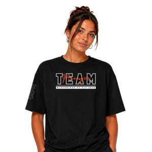 May include: A black short-sleeved t-shirt featuring the white outlined text TEAM with the red script word Manager overlaid. Below this, a white rectangular bar contains the black text BLACKRIDGE FC U12 2024. A small block of text is visible on the sleeve.
