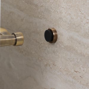 Op de afbeelding: Close-up van een bronzen deurstopper met een zwarte rubberen bumper. De deurstopper is gemonteerd op een lichtbeige getextureerde muur. Een bijpassende bronzen deurklink is zichtbaar op de achtergrond.