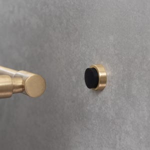 Puede incluir: Un tirador de puerta de latón y un tope de puerta a juego. El tirador de la puerta es cilíndrico con un extremo redondeado. El tope de la puerta tiene un parachoques de goma negro y está montado en una pared gris. El hardware tiene un diseño moderno y minimalista.