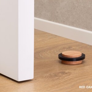 May include: A red oak door stop with a black band, 0.87 inches in diameter, rests on a wood floor. The door stop is near a white door and a wall with a white baseboard. The text "RED OAK - 22mm = 0.87" is visible.