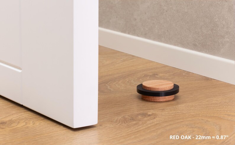 May include: A red oak door stop with a black band, 0.87 inches in diameter, rests on a wood floor. The door stop is near a white door and a wall with a white baseboard. The text "RED OAK - 22mm = 0.87" is visible.