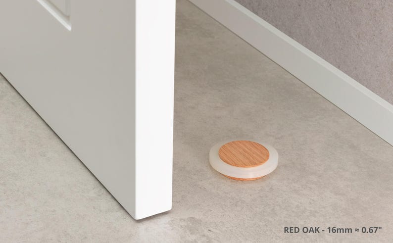 May include: A round door stop with a red oak wood center and a clear, rubbery outer ring. The door stop is 0.67 inches in diameter and is on a light gray floor. The text "RED OAK - 16mm &asymp; 0.67" is at the bottom.