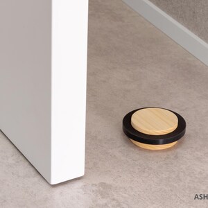 May include: A wooden door stop with a black rubber ring and a light wood top. The door stop is approximately 0.87 inches in diameter and is resting on a light-colored floor next to a white door.