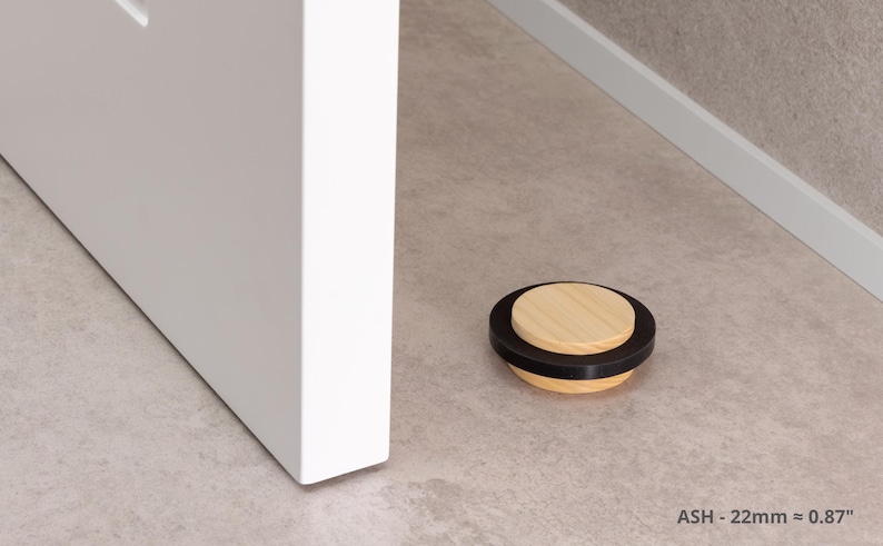 May include: A wooden door stop with a black rubber ring and a light wood top. The door stop is approximately 0.87 inches in diameter and is resting on a light-colored floor next to a white door.