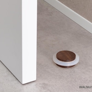 May include: A walnut door stop with a white rubber ring. The door stop is 0.87 inches in diameter and is on a light gray floor next to a white door.