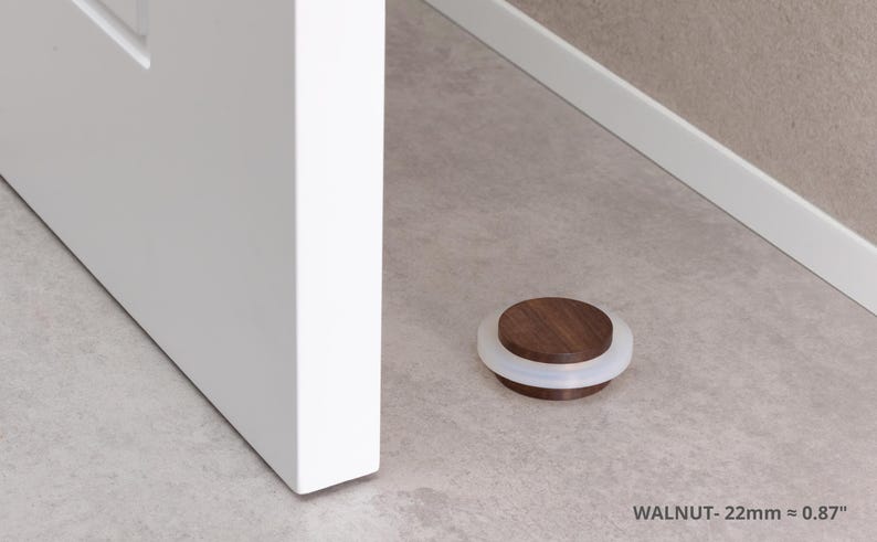May include: A walnut door stop with a white rubber ring. The door stop is 0.87 inches in diameter and is on a light gray floor next to a white door.