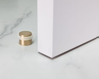 May include: A gold-colored door stop with a white rubber bumper. The cylindrical door stop is next to a white door. The door stop is a decorative and functional item for home decor.