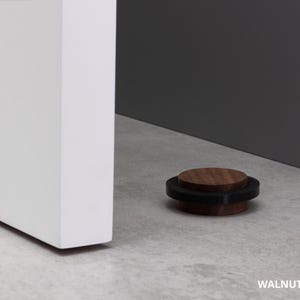 May include: A walnut door stop, 0.87 inches in diameter, with a black rubber ring. The door stop is on a light gray surface next to a white door and a gray wall. The text "WALNUT - 22mm &asymp; 0.87" is visible.