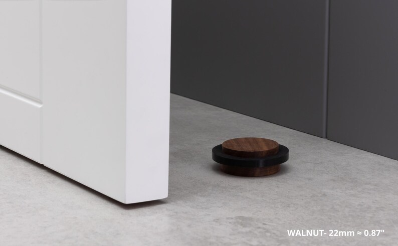 May include: A walnut door stop, 0.87 inches in diameter, with a black rubber ring. The door stop is on a light gray surface next to a white door and a gray wall. The text "WALNUT - 22mm &asymp; 0.87" is visible.