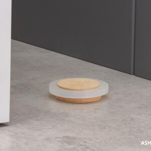 May include: A round, decorative soap dish with a wooden base, a frosted white middle section, and a light-colored wooden top. The dish is approximately 0.63 inches in height. The background is a neutral gray.