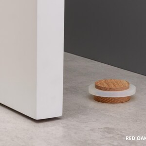 May include: A red oak door stop with a white rubber ring. The door stop is 0.87 inches in diameter and is resting on a light gray surface near a white door and a dark gray wall.