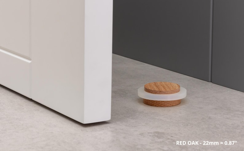 May include: A red oak door stop with a white rubber ring. The door stop is 0.87 inches in diameter and is resting on a light gray surface near a white door and a dark gray wall.