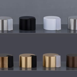 May include: A collection of cylindrical door stops in various metallic finishes, including silver, black, gold, and bronze. Each door stop has a contrasting rubber bumper. The door stops are displayed on a gray surface.