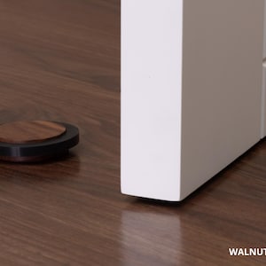 May include: A walnut door stop, 0.63 inches in diameter, rests on a dark wood floor next to a white door. The door stop has a dark rim and a circular walnut top. The text "WALNUT - 16mm = 0.63" is visible.