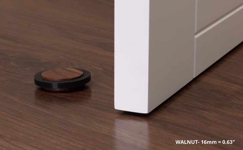May include: A walnut door stop, 0.63 inches in diameter, rests on a dark wood floor next to a white door. The door stop has a dark rim and a circular walnut top. The text "WALNUT - 16mm = 0.63" is visible.