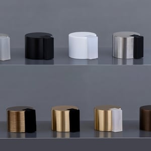 Könnte beinhalten: Eine Sammlung zylindrischer Türstopper in verschiedenen metallischen Ausführungen, darunter Silber, Schwarz, Gold und Bronze. Jeder Stopper verfügt über einen kontrastierenden Gummi- oder Kunststoffpuffer. Die Türstopper sind auf einem grauen Regal angeordnet.