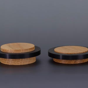 May include: Two wooden door stops with a black ring and a light brown top. The door stops have a circular design and are made of wood. The background is a solid gray color. The logo "EVI HERRAJES" is in the top right corner.