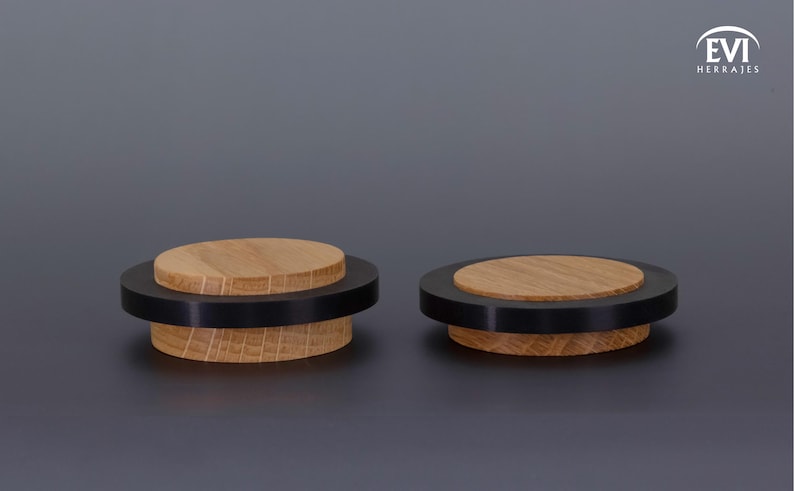 May include: Two wooden door stops with a black ring and a light brown top. The door stops have a circular design and are made of wood. The background is a solid gray color. The logo "EVI HERRAJES" is in the top right corner.