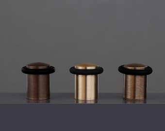 May include: Three metallic door stops with a circular top and a cylindrical base. Each door stop has a black rubber ring around the base of the top. The door stops are in shades of bronze and gold. The dimensions are 240x35mm.
