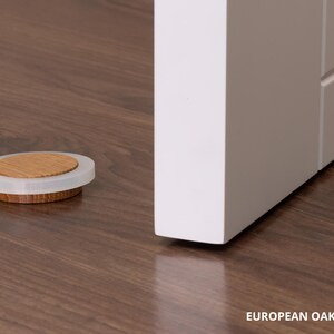 May include: A wooden and clear plastic door stopper sits on a dark brown wooden floor. The door stopper is circular with a light brown wooden center and a clear plastic rim. The text "EUROPEAN OAK - 16mm &asymp; 0.63" is at the bottom.