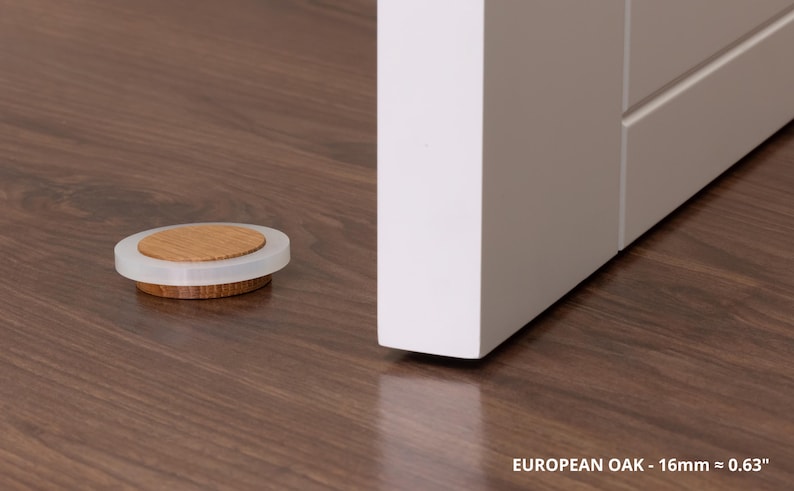 May include: A wooden and clear plastic door stopper sits on a dark brown wooden floor. The door stopper is circular with a light brown wooden center and a clear plastic rim. The text "EUROPEAN OAK - 16mm &asymp; 0.63" is at the bottom.