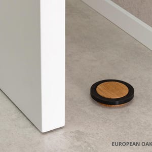 May include: A round door stop made of European oak with a black rubber ring. The door stop is 0.63 inches in diameter and is resting on a light-colored floor. A white door and trim are in the background.