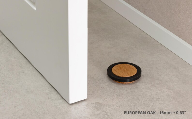 May include: A round door stop made of European oak with a black rubber ring. The door stop is 0.63 inches in diameter and is resting on a light-colored floor. A white door and trim are in the background.