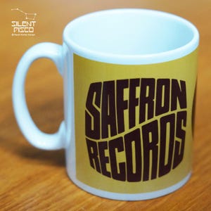May include: White ceramic mug with a yellow background and black text that reads "SAFFRON RECORDS".