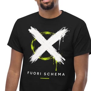 Può includere: T-shirt nero con un design a 'X' bianco su un cerchio verde neon e un effetto di vernice gocciolante. Il testo "FUORI SCHEMA" è stampato sotto. La maglietta è indossata da una persona che indossa jeans blu.