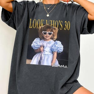 Peut inclure: Un t-shirt gris anthracite à manches courtes avec une impression graphique représentant un jeune enfant portant des lunettes de soleil en forme de cœur et une robe bleu clair. Le texte "LOOK WHO'S 30" est en doré et le nom "Aurora" en blanc.