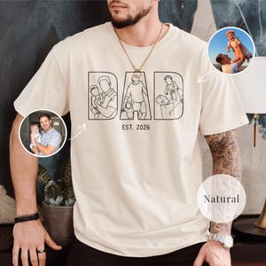 May include: A cream-coloured t-shirt featuring the word "DAD" in black line art, with "EST. 2026" below. The design shows a father with children. Includes two circular photo inserts. The shirt is a natural colour.