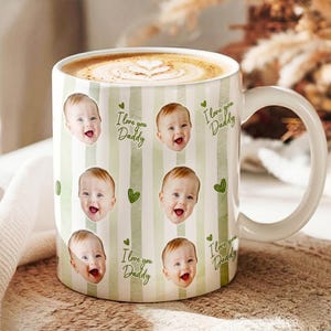 May include: White ceramic coffee mug with a handle, filled with a latte. The mug features a repeating pattern of baby faces, green hearts, and the text "I love you Daddy" on a striped background. A perfect gift for Father's Day.