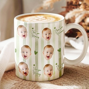 May include: White ceramic mug with a handle, filled with coffee and latte art. The mug features a green and white striped pattern with baby faces, green hearts, and the text "I love you Mommy".