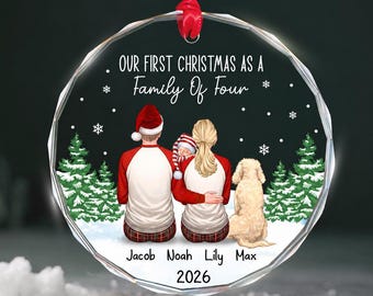 May include: A round, clear glass ornament with a red ribbon, featuring a printed illustration of two adults, a baby, and a dog in a snowy forest. The text reads: Our First Christmas As A Family Of Four, Jacob, Noah, Lily, Max, and the year 2026.