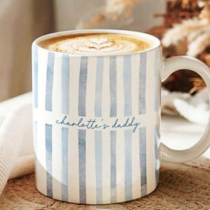 May include: A white ceramic mug with vertical blue stripes and the text "charlotte's daddy" in cursive. The mug is filled with a cappuccino, topped with a heart-shaped design in the foam. The mug has a large, curved handle.