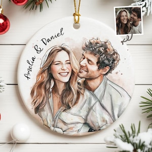 May include: A circular ceramic ornament with a watercolour illustration of a smiling couple, inscribed with the names "Amelia & Daniel". The ornament has a gold hanging string and a small photo in the upper right corner.