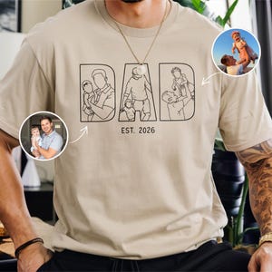 May include: A beige t-shirt with the word "DAD" in black block letters, featuring line drawings of a father with children. The text "EST. 2026" is below the word "DAD". Two circular photos are on the shirt.