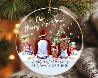 May include: A round clear Christmas ornament featuring an illustration of a family of three sitting in the snow. The text reads Our Merry Christmas, 2026, Daddy, Noah, Mommy, and AS A FAMILY OF THREE. It includes a tree, gifts, and a snowman.