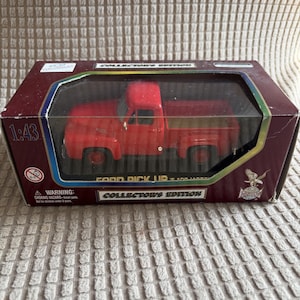 May include: A red Ford pickup truck model in a collector's edition box. The model is a 1:43 scale replica. The box is maroon with a clear plastic window, displaying the truck. The box has the text "Collector's Edition" and a warning label.