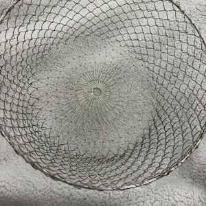 May include: A round, silver-colored wire mesh basket featuring a fine, interconnected diamond-shaped pattern. The structure is constructed from thin metal wire, forming a deep, bowl-like shape with a reinforced circular rim and a central point.
