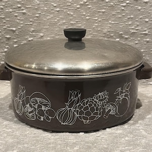 May include: A brown cooking pot with a stainless steel lid and a black handle. The pot features a white outline design of various vegetables, including mushrooms, onions, and artichokes. The pot is designed for cooking.